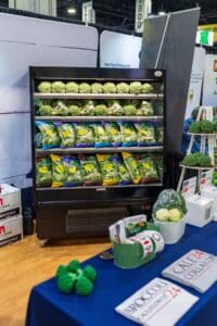 Display of bagged salads and broccoli heads in a refrigerated case at a produce booth, with promotional materials and broccoli-themed items on a nearby table.