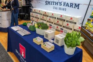 A table with promotional materials, brochures, QR code boxes, fresh broccoli, and lettuce. A "Smith's Farm Stag Brand" banner is displayed in the background.