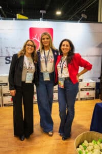 Three women stand together in front of a booth display, all wearing casual business attire and lanyards.