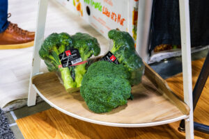 Three packaged broccoli heads on a wooden shelf, with a pair of brown shoes partially visible in the background.