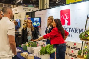 People interacting at a booth displaying farm products and promotional materials, with boxes and fresh produce visible.