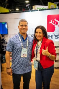 Two people smiling and posing together at an event. One person is wearing a blue shirt, and the other is in a red blazer giving a thumbs-up. Event signage and booths are visible in the background.