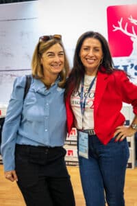 Two women smiling and posing together indoors, one wearing a blue shirt and the other a red jacket.